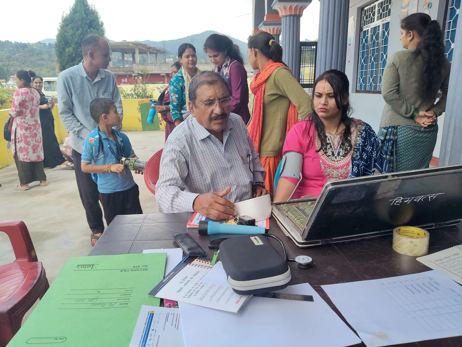 A doctor takes a woman's blood pressure at a table on a school veranda, an open trial lens set beside him and others waiting their turn.