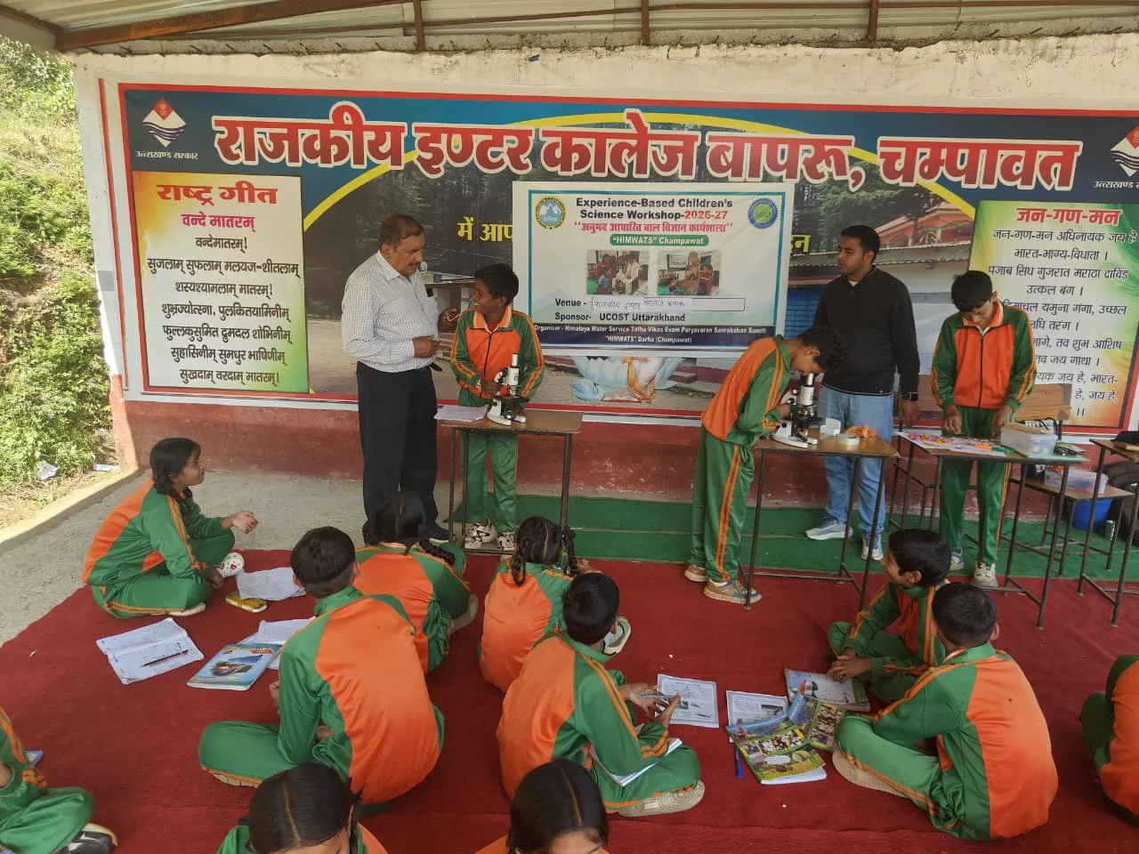 Students in green and orange tracksuits work at microscopes on small tables beneath a workshop banner, while the rest of the class sits on a red carpet with their notebooks.