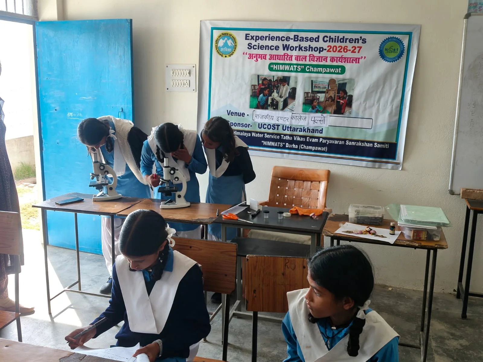Three pupils in blue-and-white uniforms look through microscopes on a classroom table beneath the workshop banner, two classmates writing in the foreground.